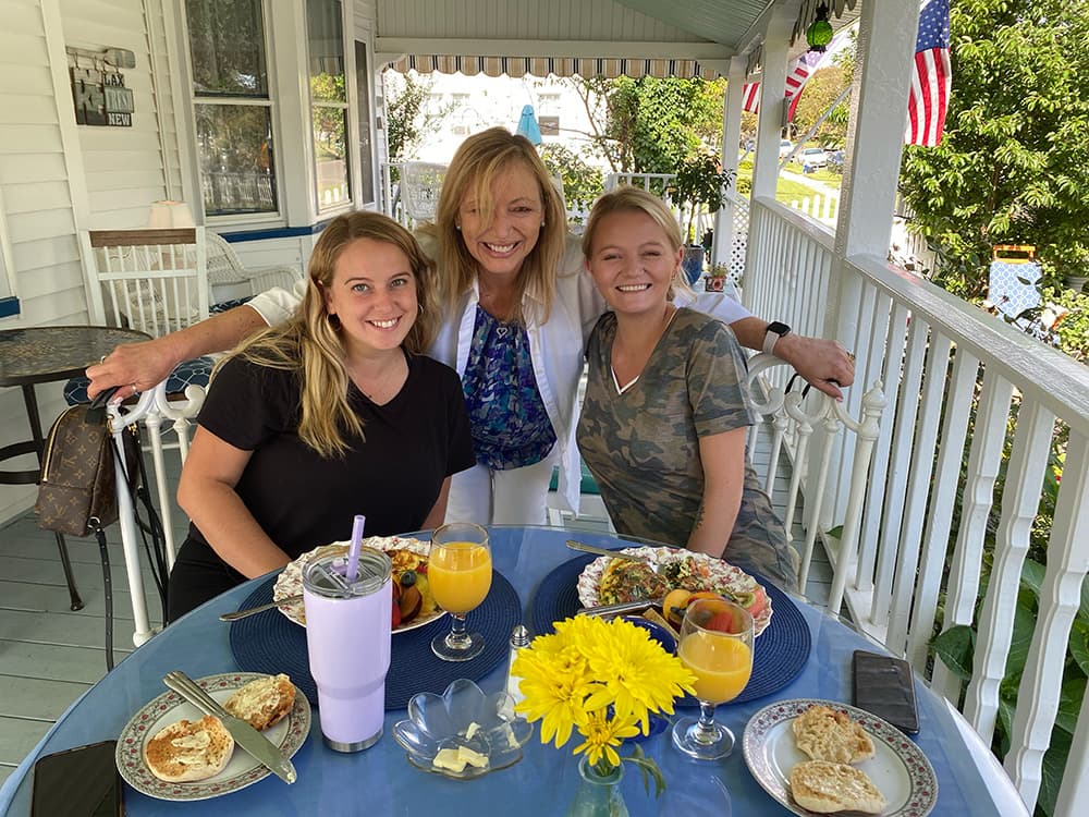 Three women smile for a photo at a table set with breakfast and drinks on a porch.