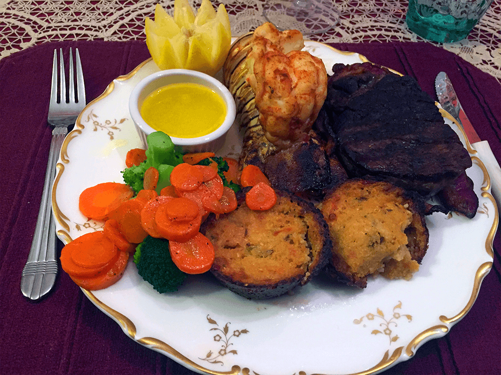 A plate featuring grilled lobster tail, steak, stuffed mushrooms, sautéed carrots, broccoli, and lemon wedges, served with a small dish of olive oil.