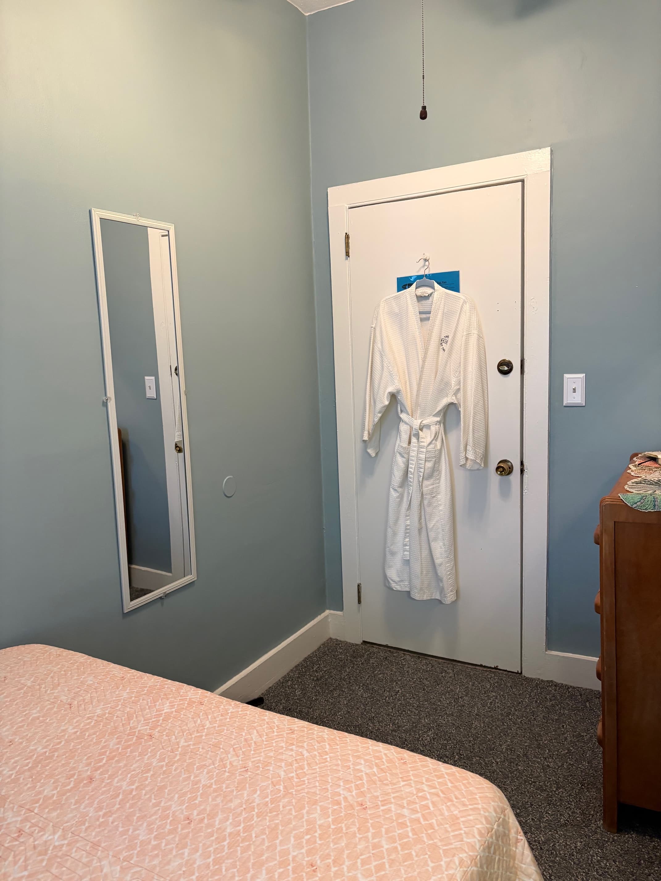 A small, light blue room featuring a bed, a mirror, and a white robe hanging on the door.