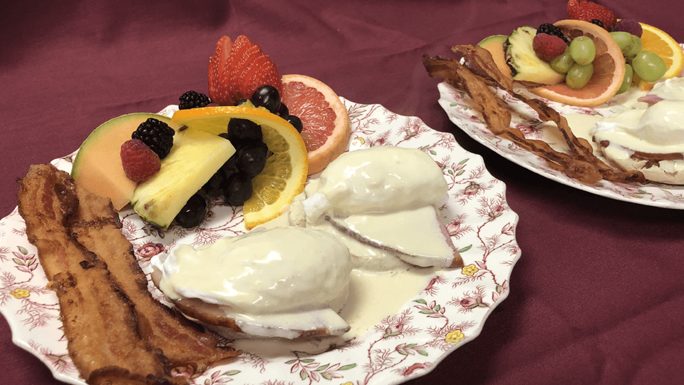 Two plates of breakfast featuring poached eggs with sauce, crispy bacon, and a variety of fresh fruit.