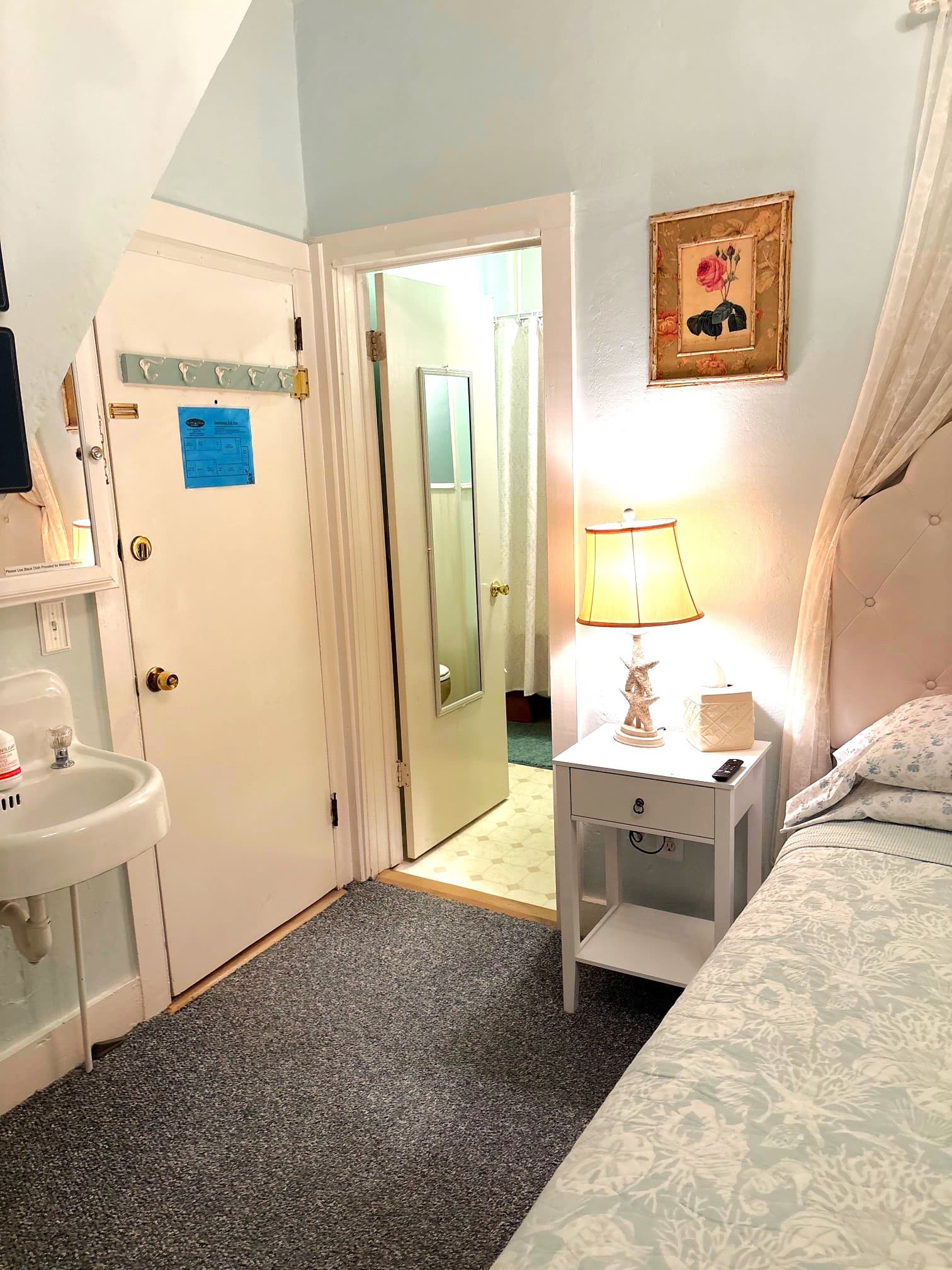 A cozy, light-colored bedroom featuring a nightstand with a lamp, a sink, and an open door leading to a bathroom.