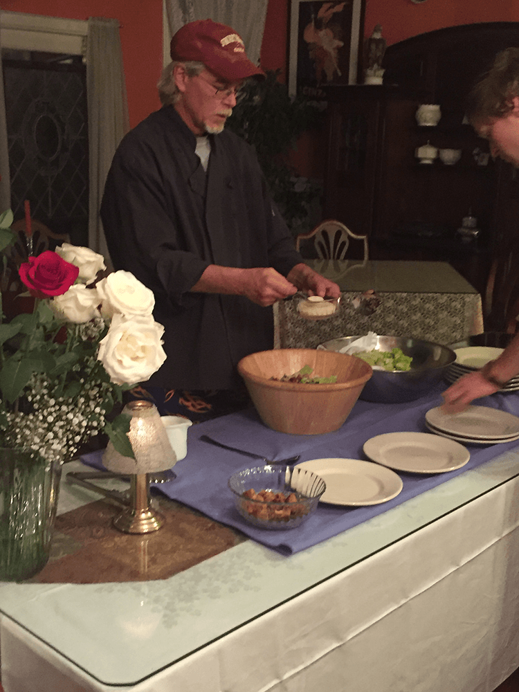 A man in a chef's coat prepares a salad at a dinner table adorned with roses and plates.