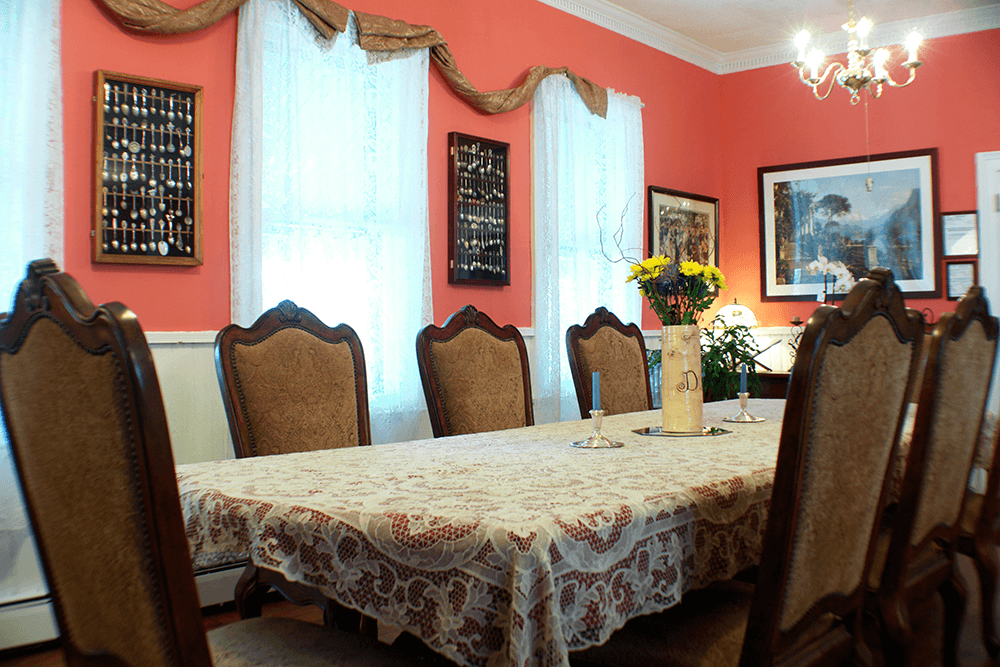 A dining room featuring a large table with a lace tablecloth, surrounded by ornate chairs and accented by colorful artwork and flowers.