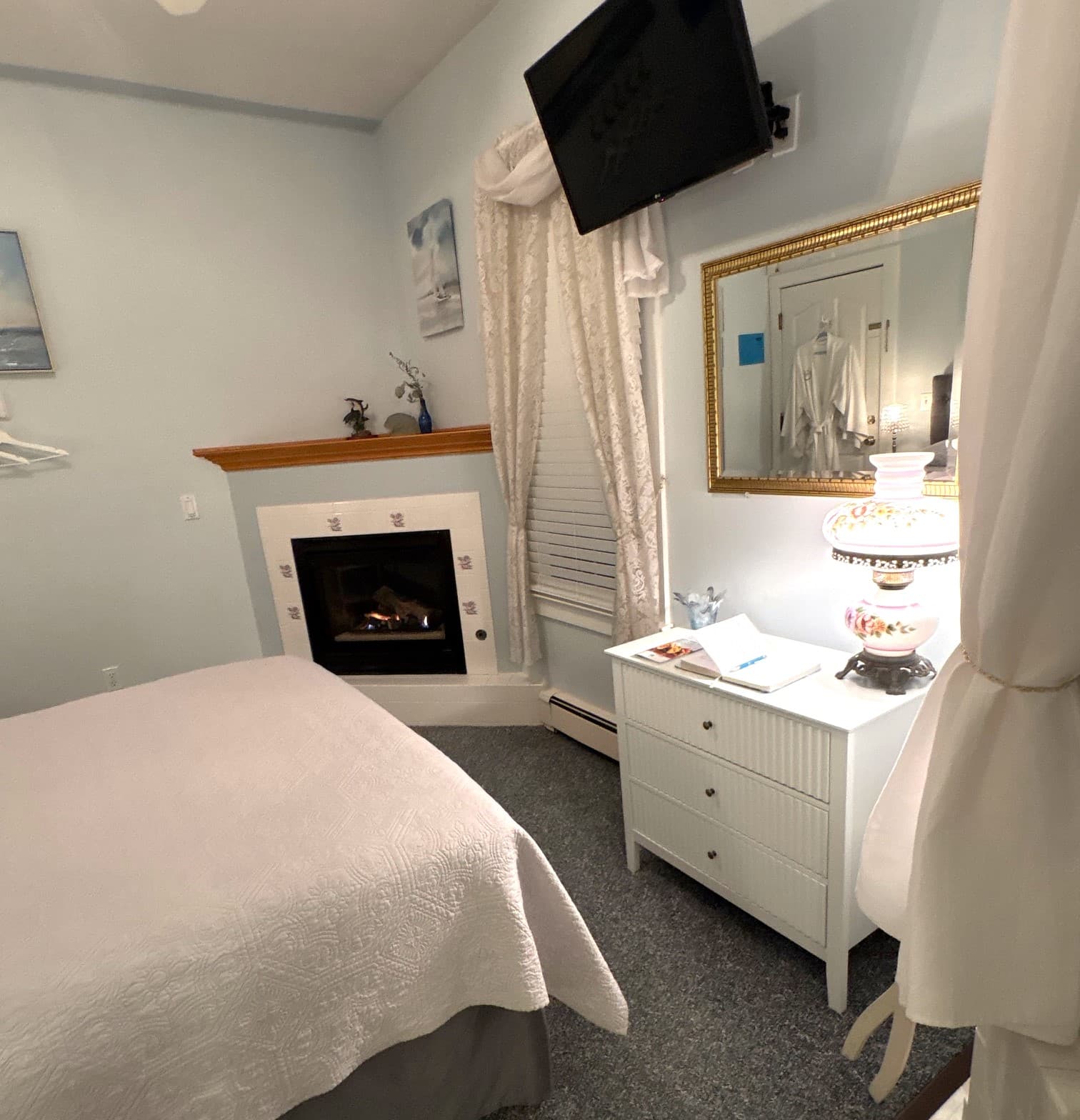 An interior view of a bright guest room featuring a plush gray tufted headboard, a bed with a circular patterned quilt, a ceiling fan, and a built-in corner fireplace with a white mantel and decorative tiles.