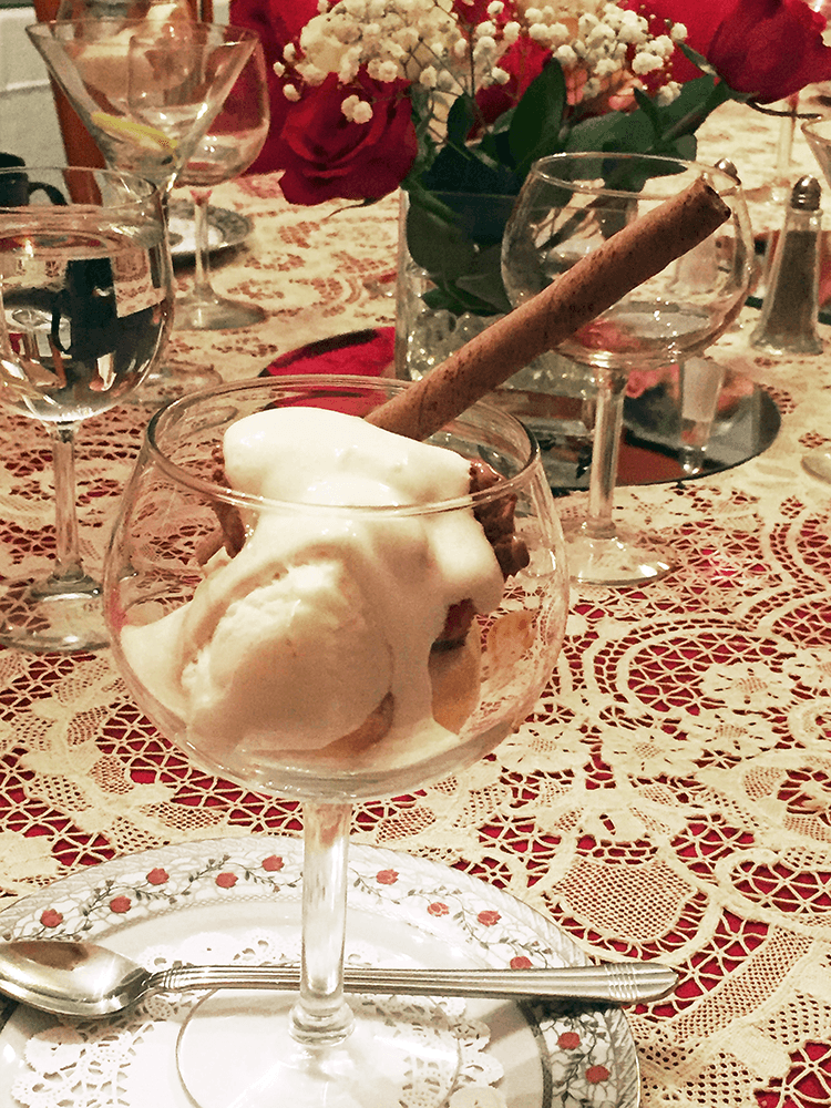 A glass dessert bowl filled with ice cream, topped with whipped cream and a cinnamon stick, amidst a table setting with roses and water glasses.